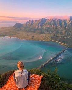 a woman sitting on top of a blanket looking out at the ocean and mountains in the distance