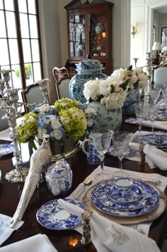 a dining room table set with blue and white plates, silverware, vases and flowers