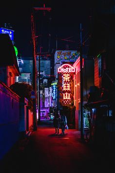 two people walking down an alley way at night with neon signs on the buildings behind them