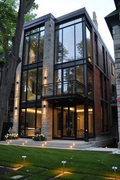 a large modern house with glass walls and lots of lights on the front porch, surrounded by lush green grass