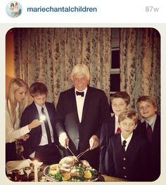 a group of people standing around a table with a cake on it and a man in a tuxedo cutting the cake