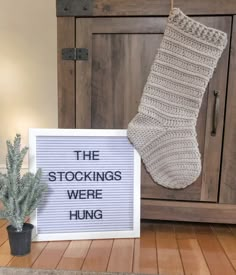 a sign that says the stockings were hung next to a potted plant and a wooden cabinet