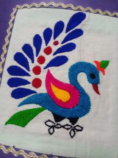 an embroidered peacock on a white cloth with blue and red accents, sitting on a purple surface