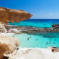 people are swimming in the clear blue water near some rocks and cliffs on a sunny day