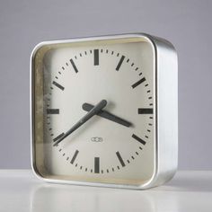 a square clock with black hands and numbers on the face is shown against a white background