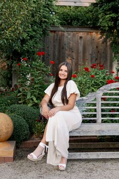 a woman sitting on a wooden bench in front of flowers and greenery, smiling at the camera