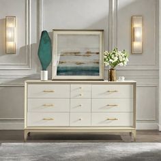 a white dresser topped with a vase filled with flowers next to two framed art pieces