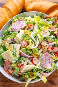 a salad in a white bowl on a wooden table next to two loaves of bread