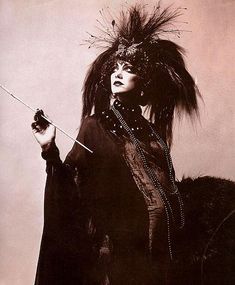 an old photo of a woman with feathers on her head, holding a stick and looking off into the distance