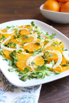 oranges and greens on a white plate next to a bowl of oranges in the background