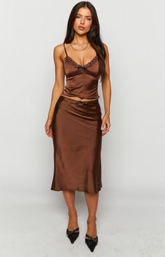 Khloe Brown Satin Midi Skirt  How to style: This sleek brown satin material gives this skirt a classic and elegant look and its minimal black lace and bow detailing is throwing us back to 2000's fashion. Pair with low brown or black heels and a matching bag.  Features:  Midi length light weight satin material Left side invisible zip Minimal lace detailing along waist Medium black ribbon bow detailing in centre Mid-rise skirt Brown Satin Midi Skirt, Satin Cami Top, Prom Midi Dress, 60's Dress, Summer Playsuit, Brown Satin, Satin Midi Skirt, Strapless Tops, Crop Top Sweater