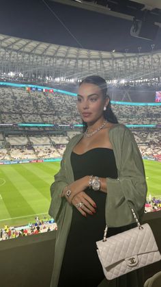 a woman standing in front of a stadium holding a white purse and looking at the camera