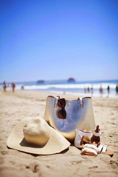 a hat, purse and shoes on the beach with people in the water behind them