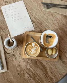 two cups of coffee on a wooden tray next to spoons and utensils