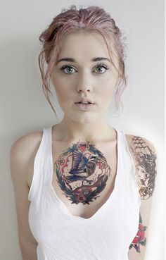 a woman with pink hair wearing a white tank top and tattoos on her chest is looking at the camera