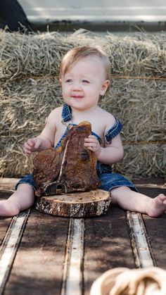 a baby sitting on the ground holding a piece of wood with hay in the background