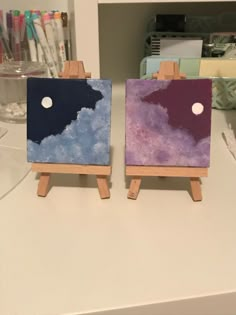 two small easels with paintings on them sitting on a table next to each other