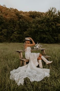 a woman sitting on a wooden bench in a field wearing a dress and cowboy hat