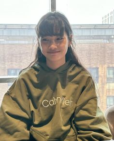 a woman sitting in front of a window wearing a green sweatshirt with the word caylen written on it