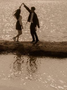 a man and woman holding hands on the shore of a body of water with sun shining in the background