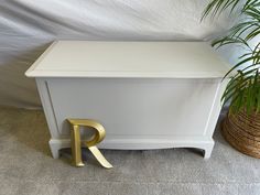 a white chest with a gold letter next to it and a potted plant on the floor