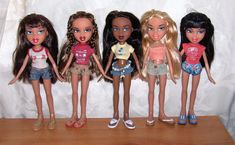 five dolls are lined up on a wooden table with white curtains behind them and one is wearing a t - shirt