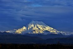 a snow covered mountain is seen in the distance with trees and clouds around it on a cloudy day