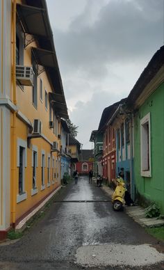 two people riding scooters down an alleyway between colorful buildings on a cloudy day