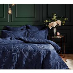 a bed with blue comforter and pillows in front of a green painted wall, next to a table with flowers