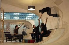 three men are sitting on the edge of a skateboard ramp and one man is jumping up into the air