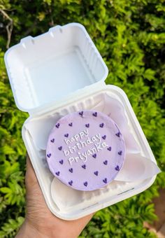 a person holding up a purple cake in a plastic container that says happy birthday priyanka