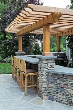 an outdoor kitchen with stone counter tops and barstools under a pergolated roof