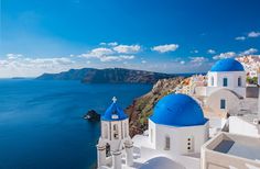 white and blue buildings on the edge of a cliff by the ocean with water in the background