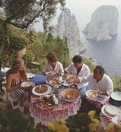 a group of people sitting around a table eating food near the ocean with cliffs in the background