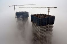 two skyscrapers in the fog with cranes on top, and one building under construction