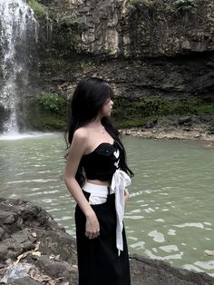 a woman standing in front of a waterfall wearing a black and white dress with a bow around her waist