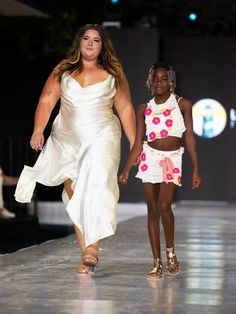 A woman in a white silk dress and a young girl in a floral-themed crochet outfit confidently walk together on a fashion runway. The background is dark, emphasizing the models and the reflective runway surface.