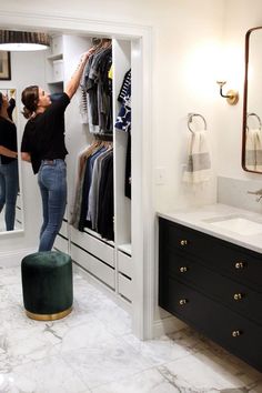 a woman hanging clothes in a closet next to a bathroom sink and vanity with mirror