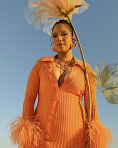 a woman in an orange dress holding a large flower and wearing feathers on her head