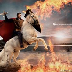 a man riding on the back of a white horse next to a fire filled field