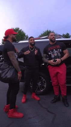 three men are standing in front of a black car and one is wearing red shoes