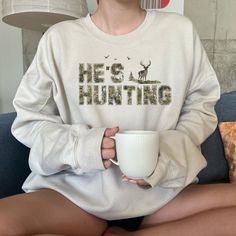 a woman sitting on a couch holding a coffee cup in her hands and wearing a sweatshirt that says he's hunting