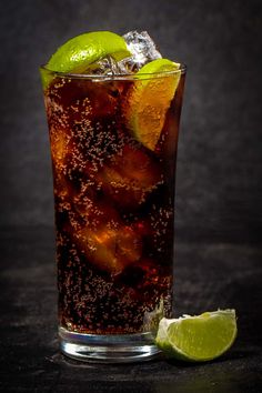 a tall glass filled with soda and limes next to a slice of lime on a black surface