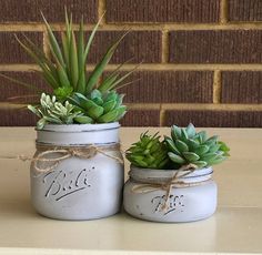 two white mason jars with succulents tied to them on top of a table