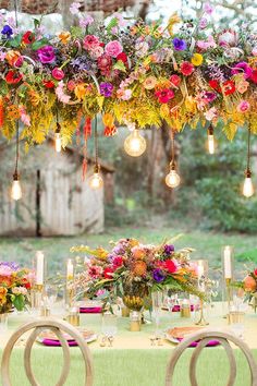 an outdoor dinner table with flowers and candles hanging from it's ceiling, surrounded by greenery