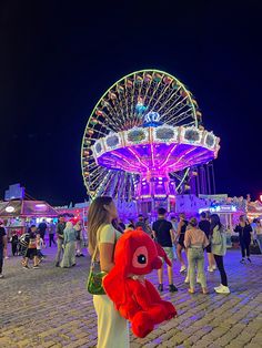 a woman holding a red stuffed animal in front of a ferris wheel at night time