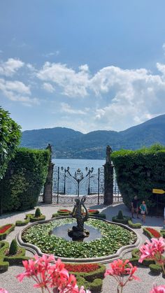 a fountain surrounded by plants and flowers in front of a body of water with mountains in the background