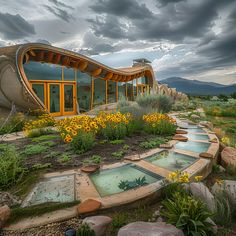 an unusual house in the middle of a field with flowers and rocks around it, under a cloudy sky