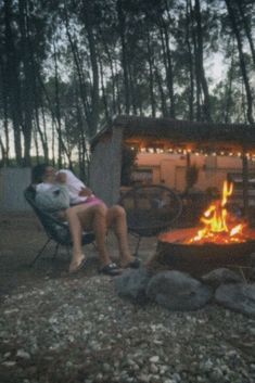 two people sitting in chairs near a fire pit with lights on the side and trees behind them