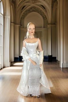 This 16-inch Robert Tonner doll from 2001 is a stunning embodiment of elegance and sophistication. Her blonde hair is styled into a sleek ponytail, showcasing her refined features. She wears classic pearl earrings and a matching pearl necklace, adding a timeless touch to her look. Her attire is truly breathtaking, featuring a beautiful silver gown that is artfully creased and ruffled, giving it a luxurious, textured appearance. The gown is accented with a bow on her left hip, adorned with crystal sequins that catch the light and add a touch of sparkle. Draped over her shoulders is a silver shawl that complements the gown perfectly, adding an extra layer of elegance. She completes her look with black high heels, which contrast beautifully with her silver ensemble. Her makeup is flawless, wi Silver Shawl, Classic Pearl Earrings, Silver Gown, Brown Lipstick, Brown Eyeshadow, Sleek Ponytail, Black High Heels, Timeless Beauty, Art Dolls